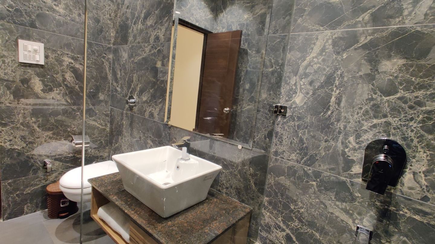 A bathroom at Hotel Exotic Amritsar featuring a textured grey tiled wall, a modern square vessel sink on a wooden vanity, a large mirror, and a black hairdryer mounted on the wall.
