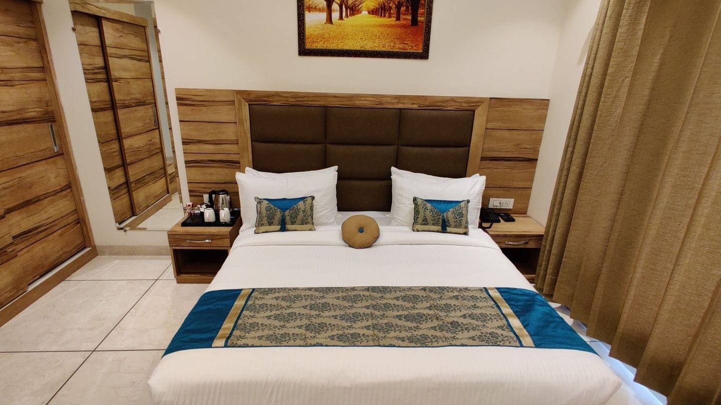  A hotel room at Hotel Exotic Amritsar featuring a king size bed with a white duvet and a patterned blue and gold throw blanket, a padded headboard, and wooden furniture including a dresser with a mirror and a bedside table. 