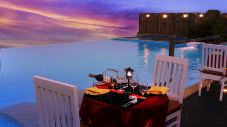 Poolside dining table at Polo Resort Cherrapunji set for two with wine, candles and yellow napkins on a deck beside the infinity pool at twilight.