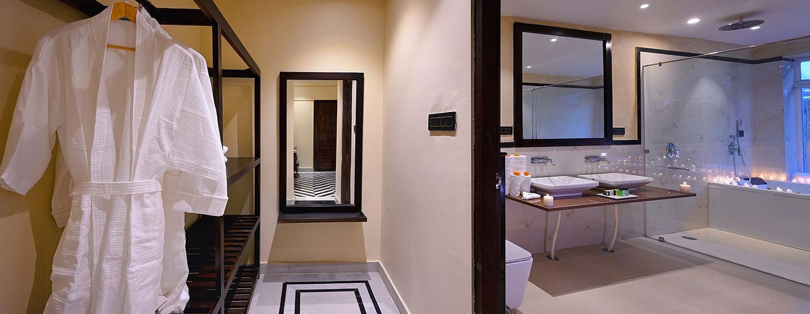 Luxury Suite Cottage entryway leading to a bathroom with double sinks, a large mirror, and a candlelit bathtub; a white bathrobe is hanging in the foreground - Singhvilas, Ranthambore