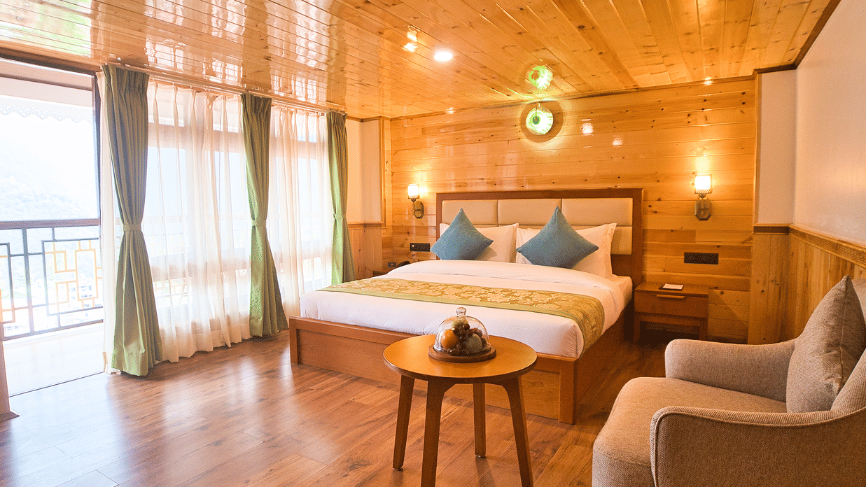 Well-lit wooden-finished room at Voyage Songfum Retreat featuring a queen bed with teal cushions, cozy seating area, and private balcony with natural light. 
