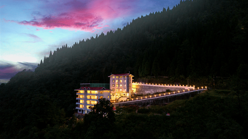 Exterior night view of Voyage Glenz Resort illuminated against a dramatic pink and blue sunset sky, surrounded by dense forest hills.