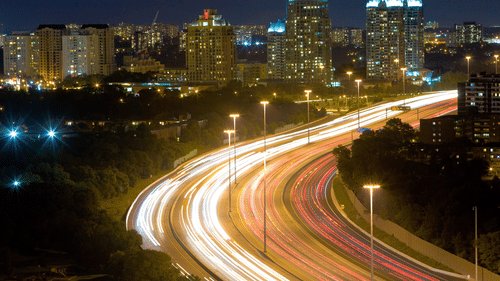 A view of a time lapse of car's headlights and taillights on a highway with buildings on the left hand side of the highway.