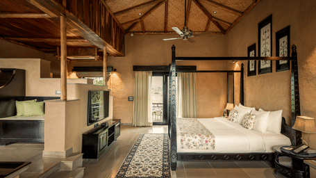 The spacious Elegant Suite at AamaGhati Wildlife Resort featuring a thatched bamboo ceiling, dark wooden bed, split-level living space with a TV console, and access to a outdoor balcony.