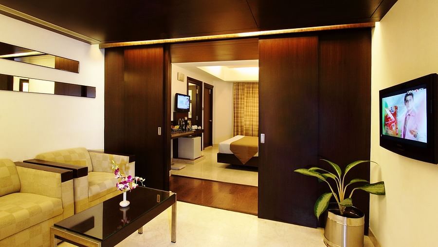 Modern hotel suite with sleek furnishings, a flat-screen TV, and a peek into the adjoining bedroom, blending comfort with style - The President Hotel Jayanagar, Bangalore