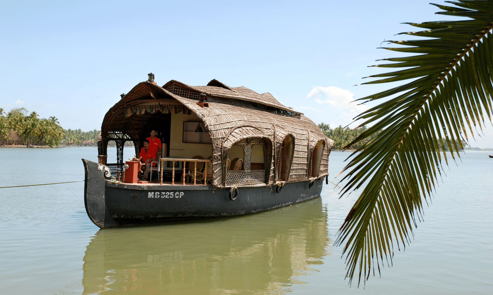 Traditional houseboat with a thatched roof floating on the water, partially framed by palm leaves at Paradise Lagoon Resort, Udupi.