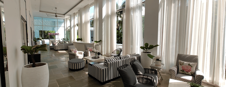 Bright hallway at The Mansion House resort with grey armchairs, striped ottomans, white conical planters, floor-to-ceiling sheer curtains, and polished light stone floor