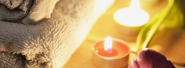 Spa setting with rolled towels, lit candles, flowers, and wooden surface, creating a relaxing atmosphere.