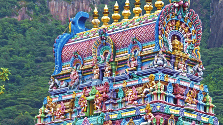 A tall multicoloured temple tower with layered carvings and statues, surrounded by greenery and hills in the background, showing detailed architecture.