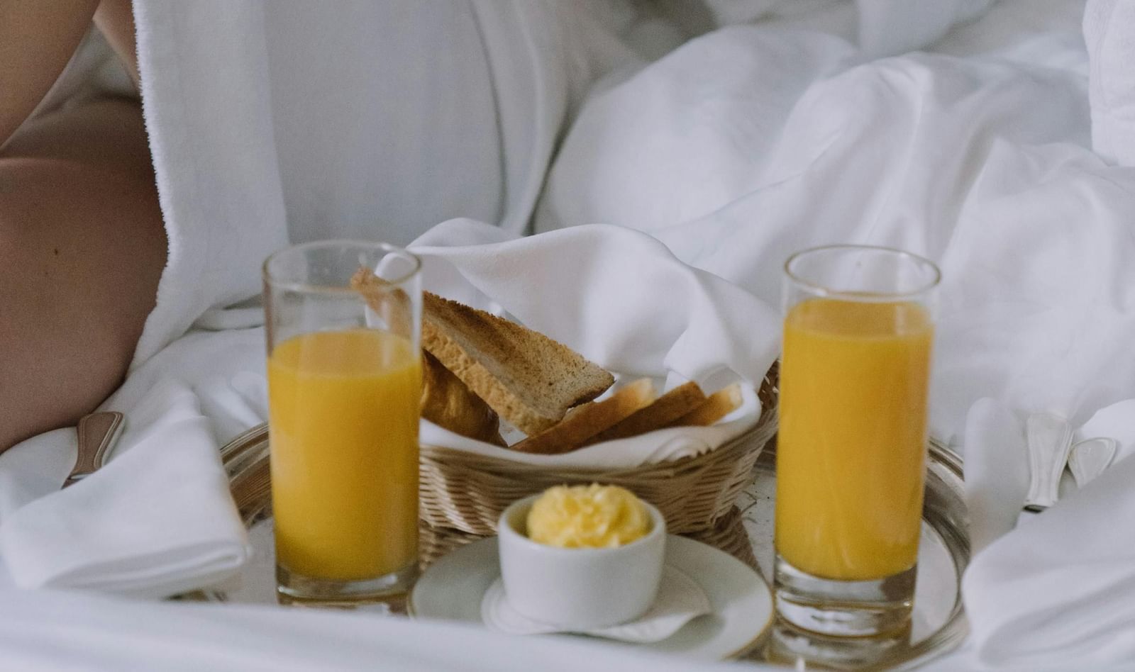A person in a white robe enjoys breakfast in bed with orange juice, toast, butter, and a plate of food on a white sheet.