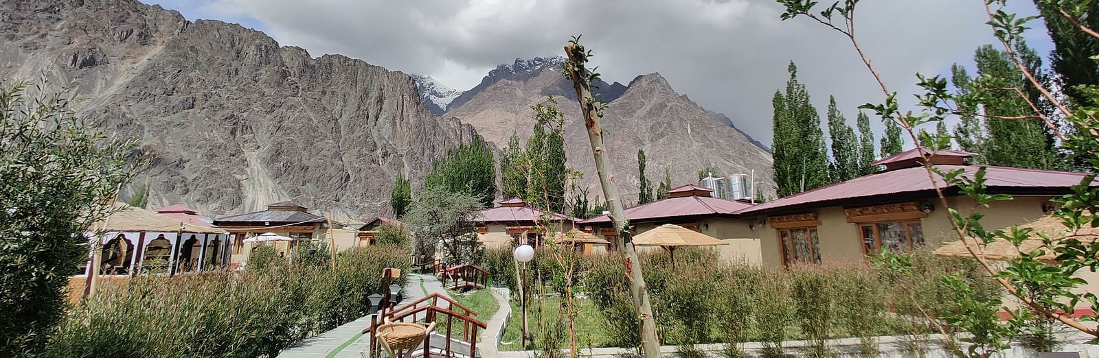A pathway leading through White Sands Resort, Ladakh, with accommodation buildings and trees on either side, set against a backdrop of mountains and a partly cloudy sky.