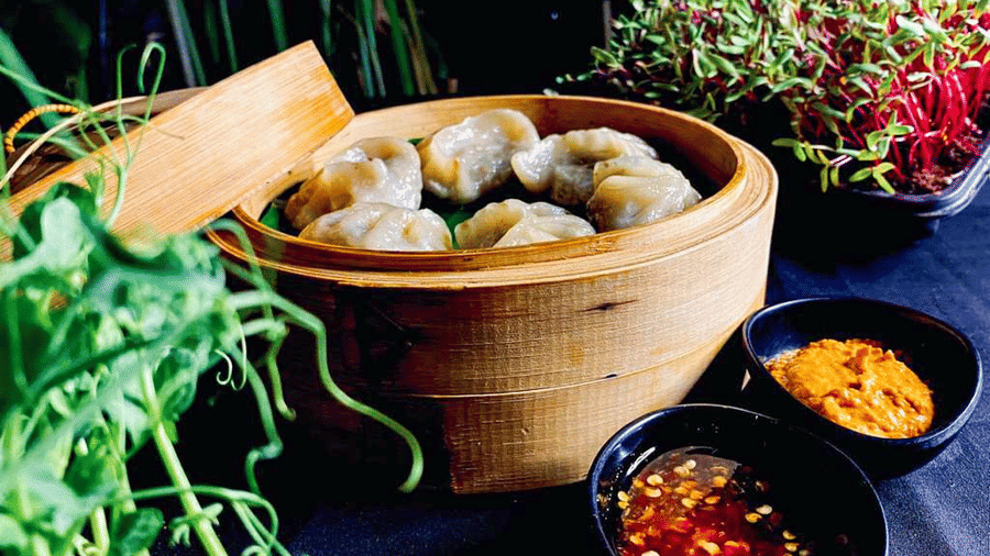 Bamboo steamer of vegetable dimsums beside 2 black bowls of orange and red chilli dipping sauces, with micro greens in the background.