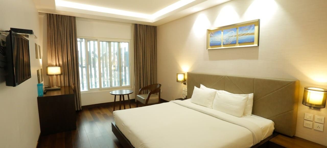 Spacious room with a large double bed, bedside lamps, wall art above the headboard, and a window with curtains pulled to the side at The Hamuse Hotel, Kodaikanal.