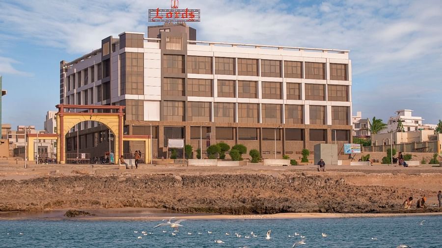 A modern multi-story hotel building with a prominent sign stands near a rocky shoreline overlooking a calm blue sea under a partly cloudy sky.