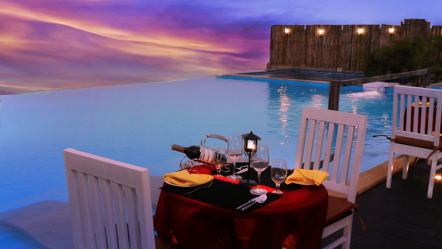 Romantic candlelit table for two by the infinity pool at Polo Resort Cherrapunji with wine, yellow napkins and pink twilight sky overhead.