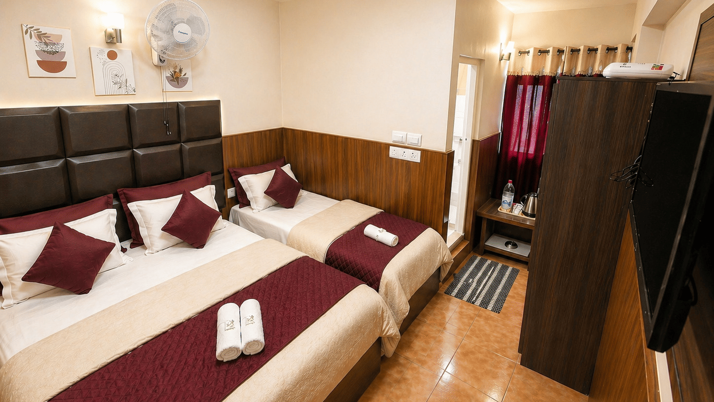 A double bed and a single bed arranged together with folded towels, a wardrobe, and a television in the Deluxe Triple Room at BN Tourist Home, Ooty.