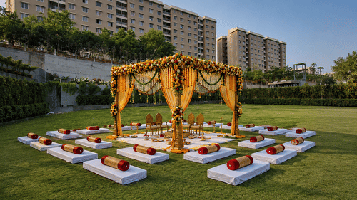 A decorated mandap canopy stands on a green lawn with floor seating cushions, bolsters, and residential apartments behind at Pride Plaza Indore.