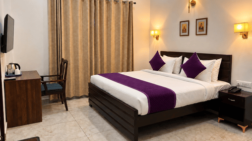 Well-appointed hotel room featuring a king size bed with purple and white linens, bedside tables, warm lighting, and beige curtains.