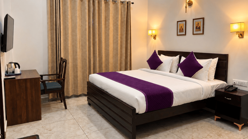 Well-appointed hotel room featuring a king size bed with purple and white linens, bedside tables, warm lighting, and beige curtains.