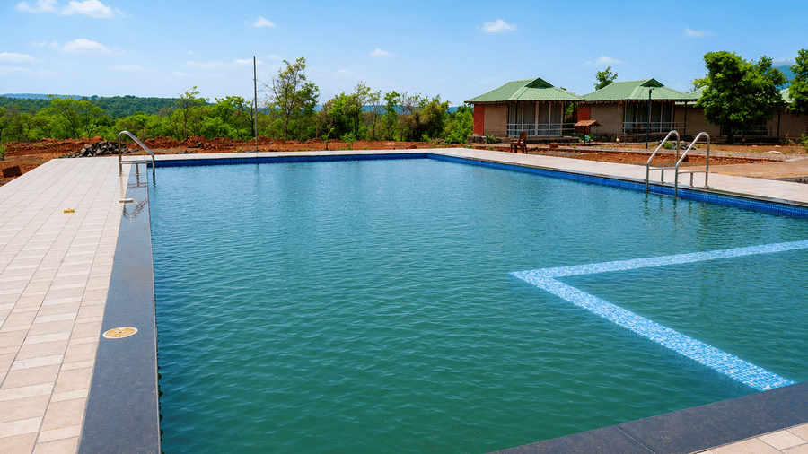 Outdoor pool at Nature Trails Kundlaika with turquoise water, granite coping, and green cottage views under a clear blue sky.