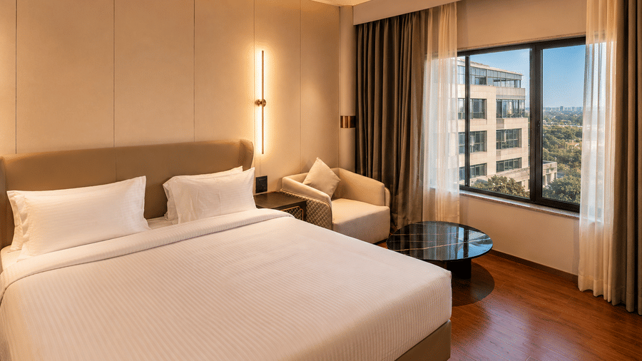 A double bed inside a Premium Room at Pride Plaza Indore, flanked by bedside lights, an armchair, a round coffee table, and a large window.