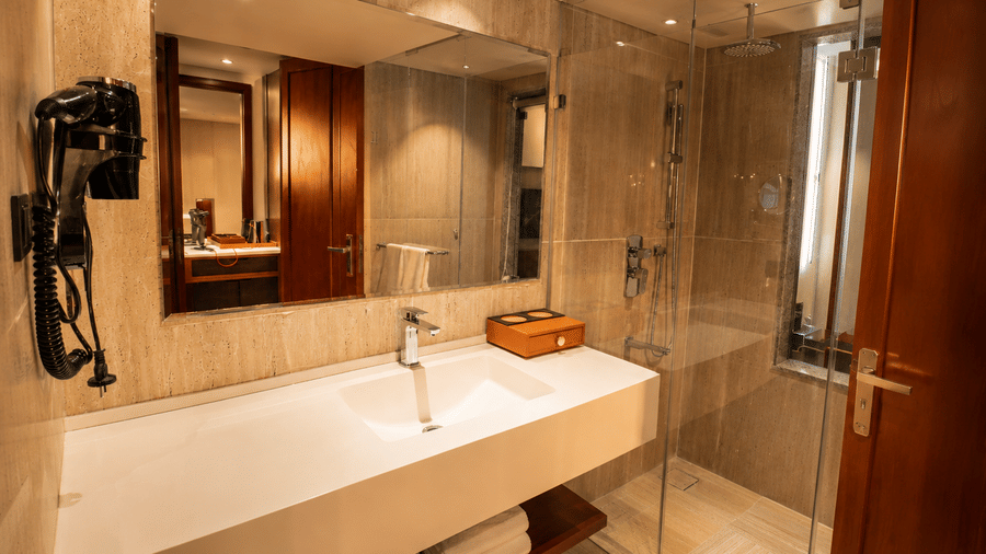 An ensuite bathroom in a Premium Room at Pride Plaza Indore with a long vanity counter, washbasin, mirror, wall hairdryer, and glass shower cubicle.