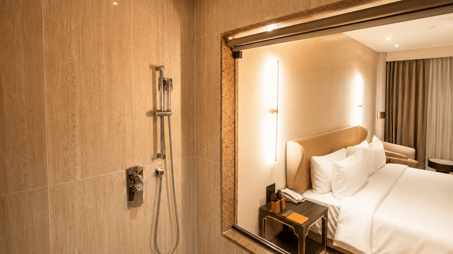 A shower fixture in the bathroom of a Premium Room at Pride Plaza Indore, with a reflection of the bedroom visible through a interior glass window.
