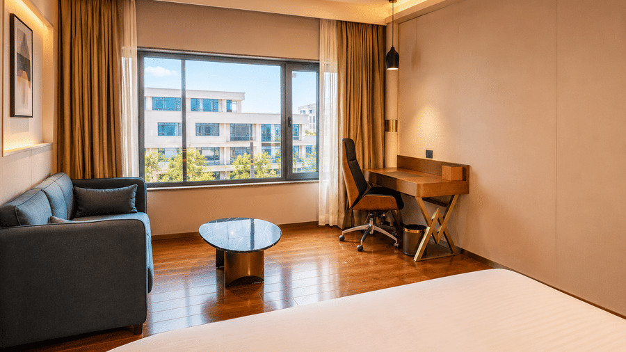 A corner of the Superior Twin Room at Pride Plaza Indore with a view of a corner of a bed, a sofa, and a desk positioned beside a window.