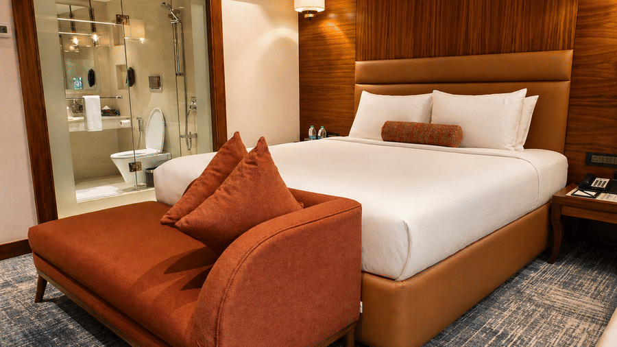Bedroom at Pride Premier Pune with a king size bed, cushioned bench and glass partition to the bathroom.