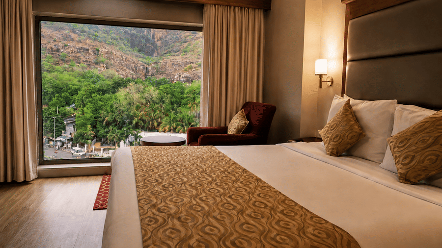 A room with a large bed, spreads, pillows, and a window with plants in the backdrop. | Raj Park Hotel - Tirupati