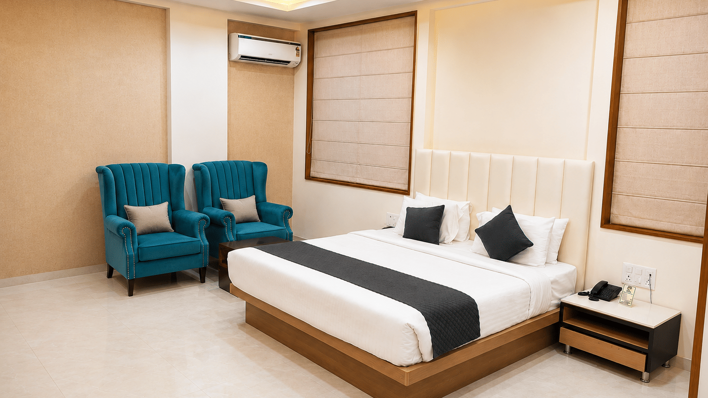 Suite at TGI Apple Inn, Ajmer Road, Jaipur featuring a bed with pillows, two armchairs, blinds on the windows, an air conditioner, and a bedside table.