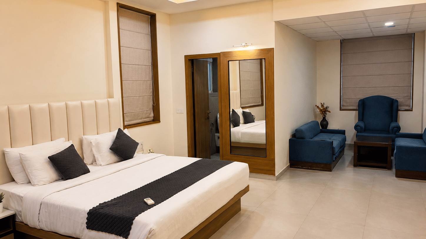 Suite at TGI Apple Inn, Ajmer Road, Jaipur featuring a bed with pillows, a seating area with armchairs and a table, blinds on the windows, and a doorway.