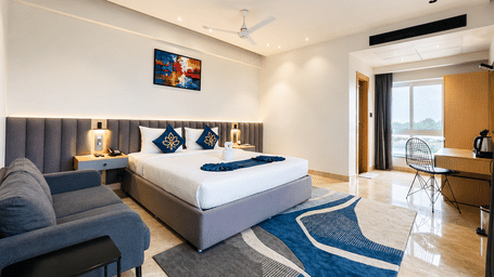 Deluxe room at Hotel Kyzen HiTech City featuring a king bed with blue accents, sofa seating, blue patterned carpet, ceiling fan, and large windows with natural light.