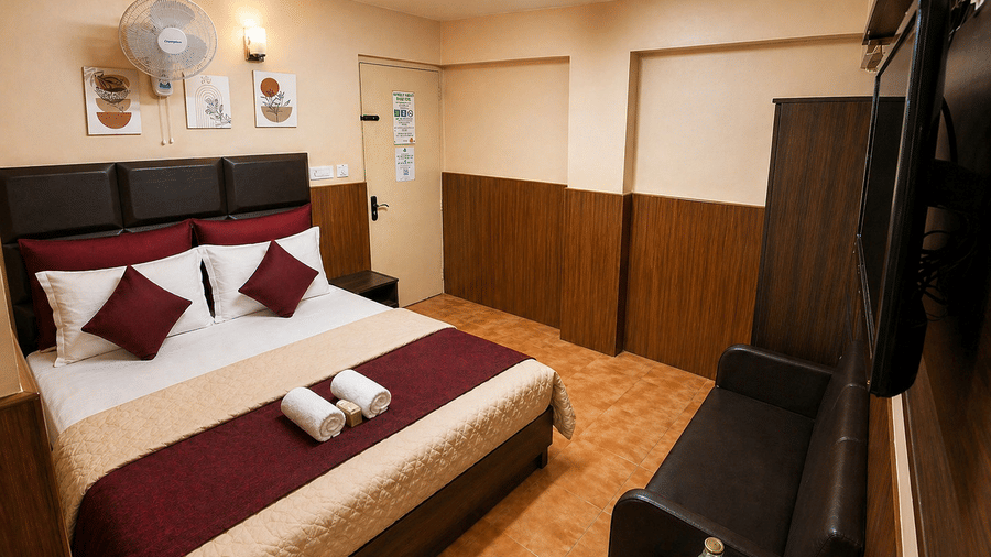 A neatly-made bed, a leather sofa, and a wall-mounted television across from the bed in the Deluxe Double Room With Extra Cot at BN Tourist Home, Ooty.