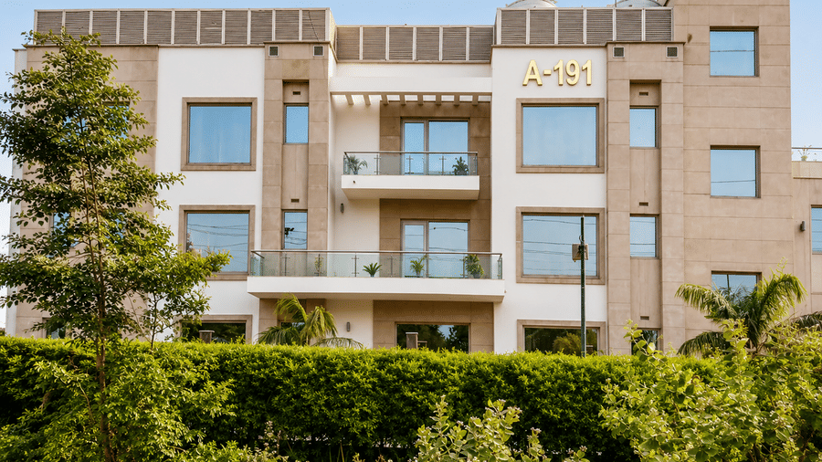 Hotel building exterior in daylight with glass balconies, landscaped hedges and tall trees set against a clear blue sky overhead above it.