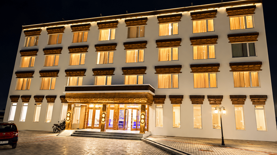 Hotel building exterior at night with illuminated windows, a lit-up carved wooden entrance canopy and a driveway with parked cars nearby.