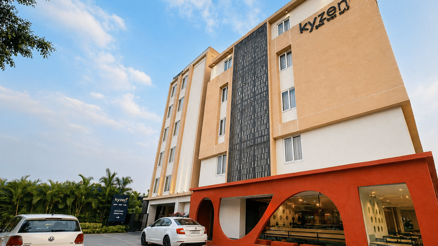 Front exterior view of Hotel Kyzen HiTech City featuring a modern building facade with orange and grey panels the hotel signage parked cars and lush green trees in the foreground.