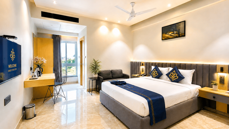 A full view of a hotel room at Hotel Kyzen HiTech City featuring a king bed with blue accent bedding grey headboard yellow accent wall work desk large windows and light tiled flooring.
