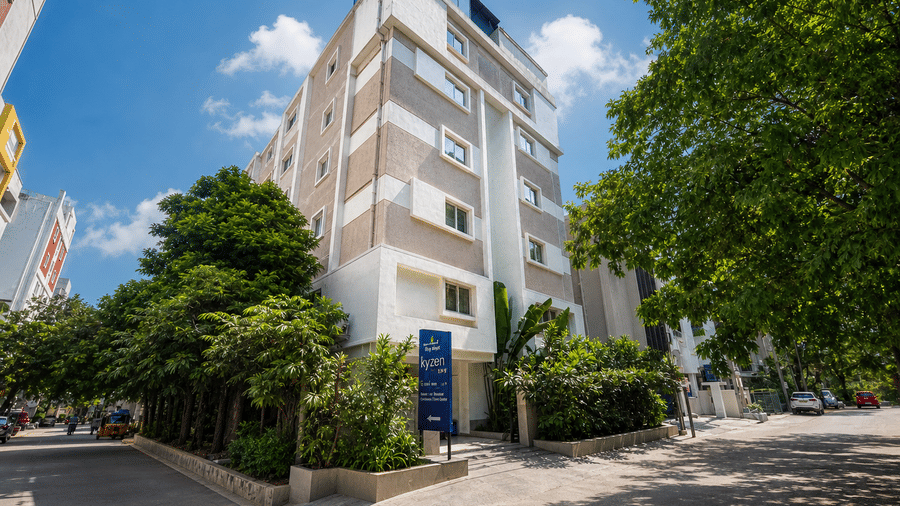  Exterior view of Hotel Kyzen Smart Stays Madhapur showing a multi-storey white building surrounded by trees under a clear blue sky.