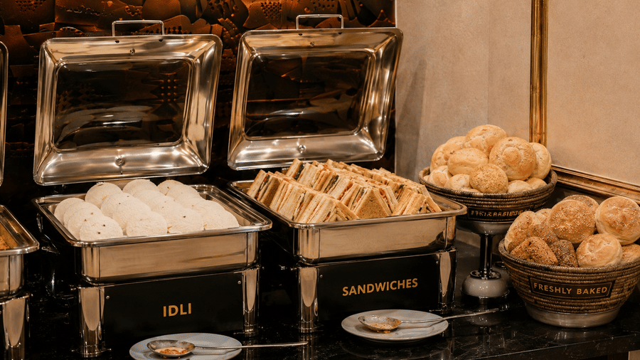 Food buffet at Lime Tree Hotel & Banquet, Jaipur, with metal chafing dishes holding idli and sandwiches alongside baskets of bread rolls.