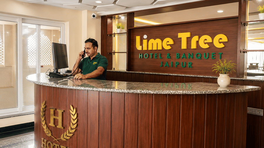 A receptionist talking on the phone behind the wooden front desk inside the reception lobby at Lime Tree Hotel & Banquet, Jaipur.