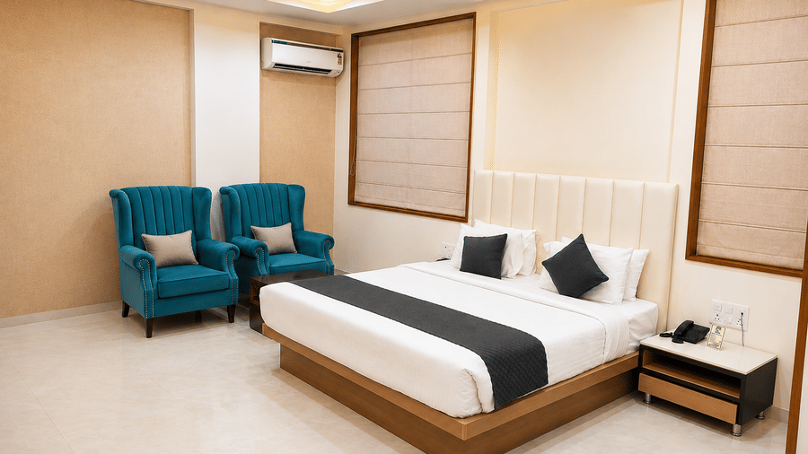 Suite at TGI Apple Inn, Ajmer Road, Jaipur featuring a bed with pillows, two armchairs, blinds on the windows, an air conditioner, and a bedside table.