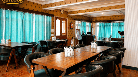 An indoor dining area featuring multiple wooden tables and upholstered chairs, with large windows in the background at Hotel Outlook, Pahalgam.