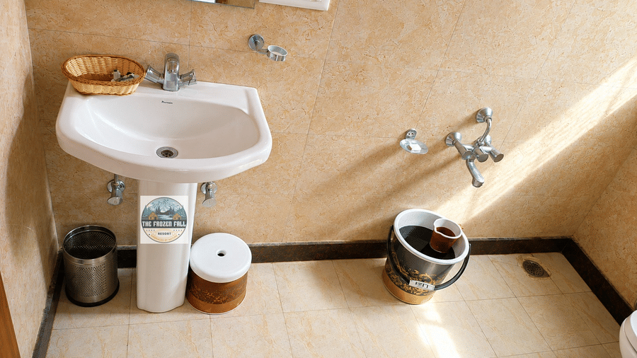 A bathroom area at Dawat e Drung by FrozenFall Resort, Drung, Tangmarg, featuring a wall-hung sink basin, a water bucket and tiled flooring.