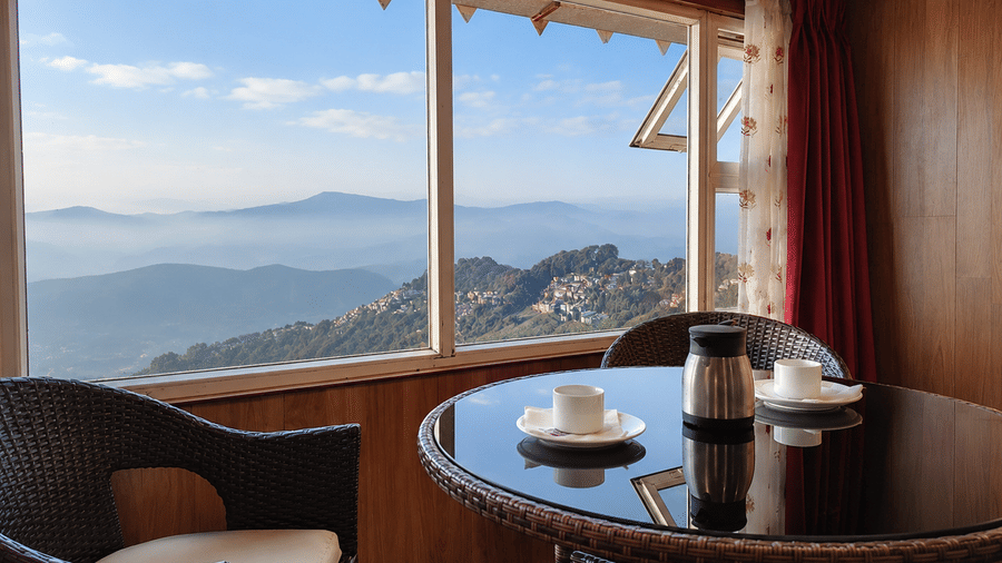 Seating nook at Valmere Thistle Boutique Hotel, Darjeeling with wicker chairs, a round table, and a panoramic view of a hillside town and mountains.