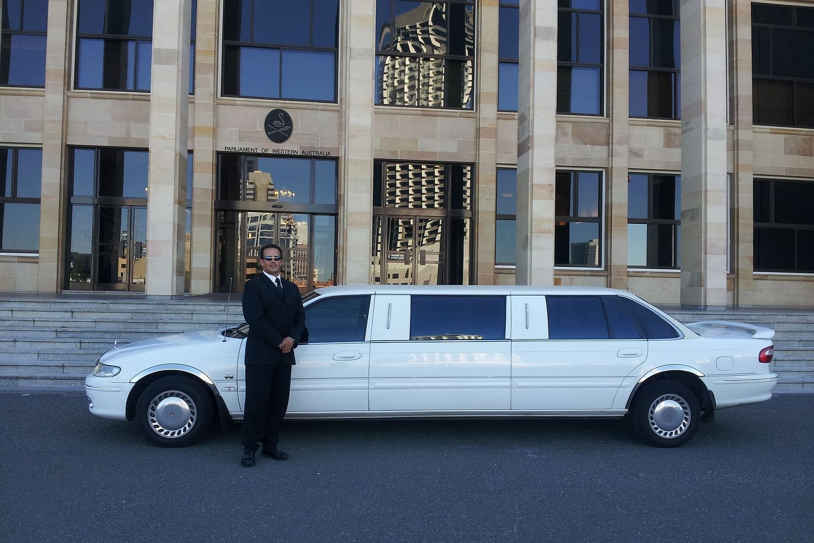 A Chauffeur standing in front of a white limousine on the road next to a building.