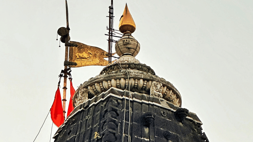 A tall temple tower with a flag and small structure on top, viewed from below against a clear sky.
