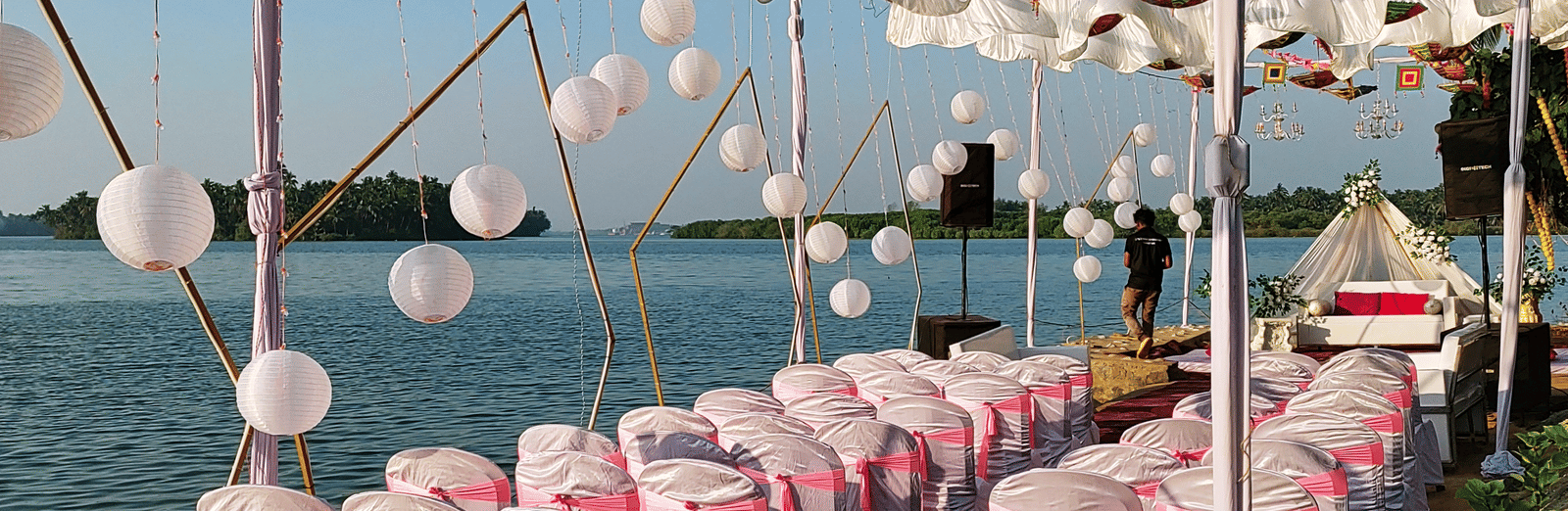 Rows of white chairs with pink covers arranged outdoors under hanging umbrellas at Paradise Lagoon Resort, Udupi.