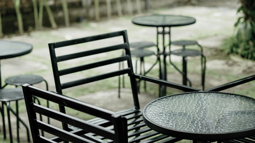 A small outdoor cafe area with metal chairs and round glass-topped tables set on a patio surrounded by dense tropical foliage.