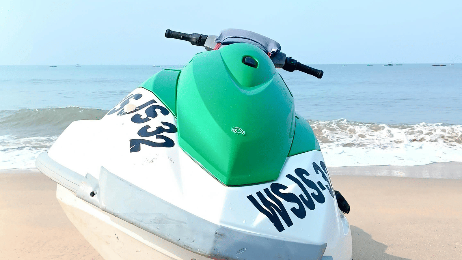  A white and green jet ski is parked on a sandy beach, with the gentle waves of the ocean and a clear blue sky in the background.